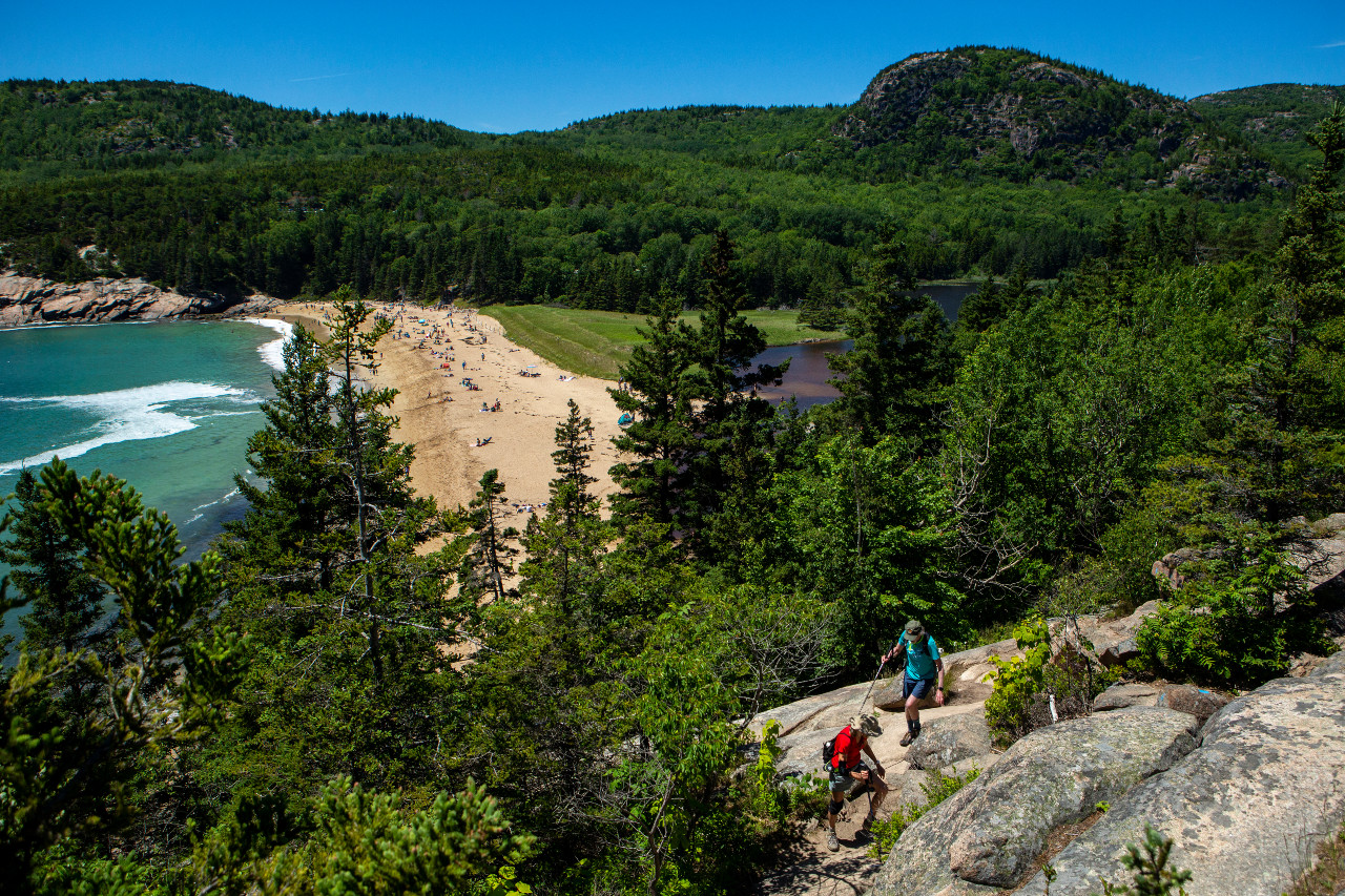 Two hikers in foreground on rocky trail overlooking a crowded beach below on a sunny spring day