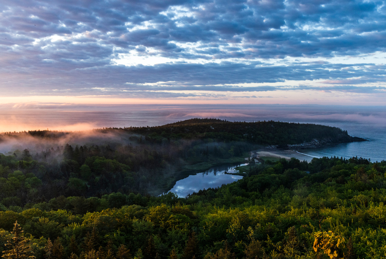 The sun rises on the horizon with tree-covered mountains and calm pond in the foreground