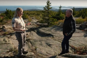 Two people stand talking on a granite summit overlooking the ocean