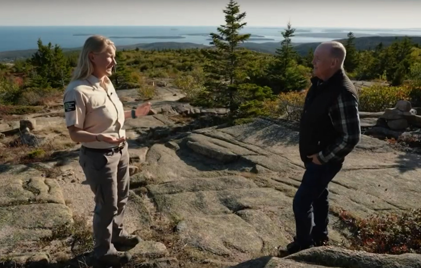 Two people stand talking on a granite summit overlooking the ocean
